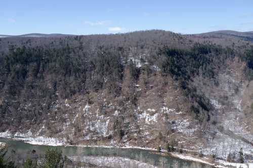 A scenic view of a snow-covered landscape with a winding river and forested hills under a clear blue sky.