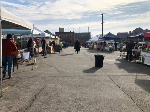 A bustling outdoor market with various vendor tents set up along a paved area under a clear sky.