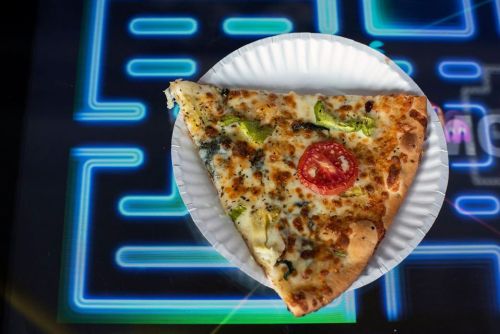 A slice of pizza with cheese, broccoli, and a tomato on a white plate, set against a colorful arcade game background.