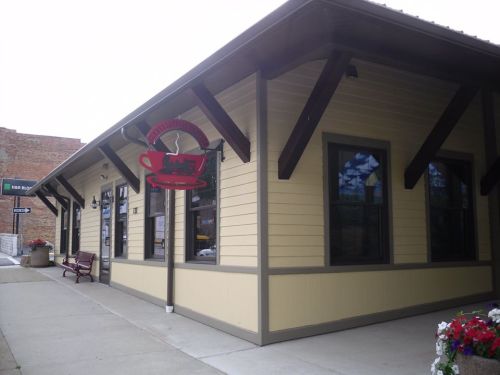 A corner café building with a red coffee cup sign, featuring large windows and a flower bed in front.