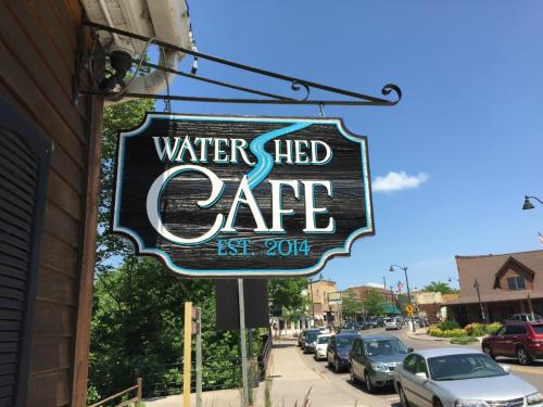 Sign for Watershed Café, established in 2014, hanging outside with a clear blue sky and street view in the background.
