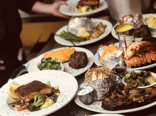 A variety of plated meals featuring salmon, steak, lobster, baked potatoes, and vegetables on a restaurant table.