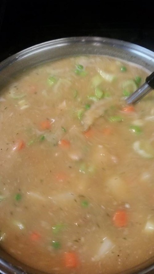 A pot of simmering soup with vegetables like carrots and celery, stirring with a spoon.