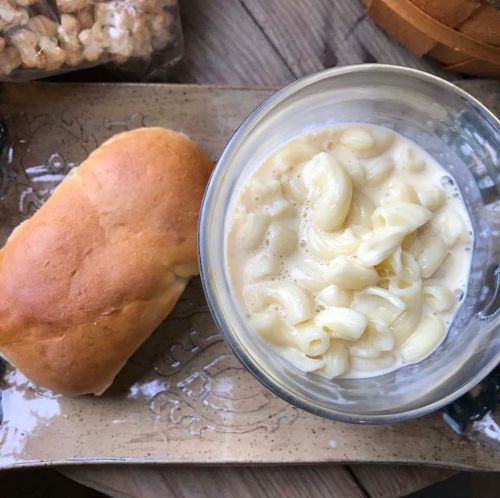 A glass bowl of macaroni and cheese next to a soft bread roll on a decorative plate.