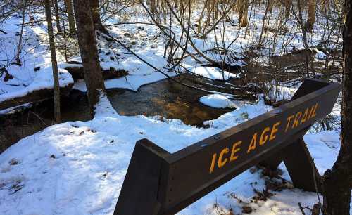 A wooden sign reading "Ice Age Trail" near a snowy landscape with a stream and trees in the background.