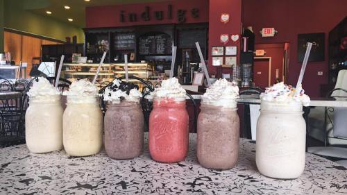 Six colorful milkshakes in mason jars topped with whipped cream, lined up on a table in a cozy café setting.