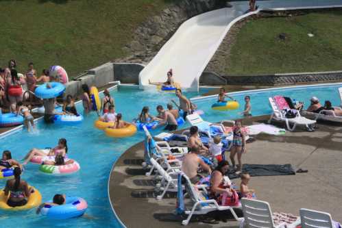 A busy water park scene with people on floats in a pool, lounging on chairs, and a water slide in the background.