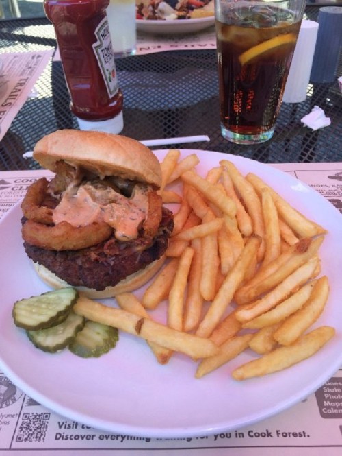 A burger topped with onion rings and sauce, served with fries and pickles, alongside a drink with a lemon slice.