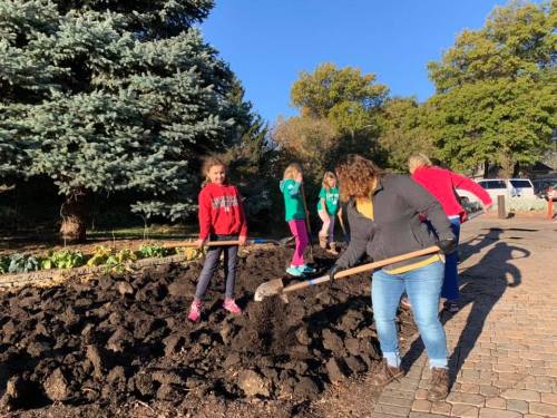 A group of people, including children, are gardening and digging in a sunny outdoor area with trees in the background.