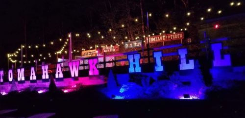 Colorful illuminated sign reading "TOMAHAWK HILL" with festive lights and trees in the background at night.