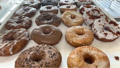 A variety of donuts on a tray, featuring chocolate, glazed, and topped with sprinkles and bacon bits.
