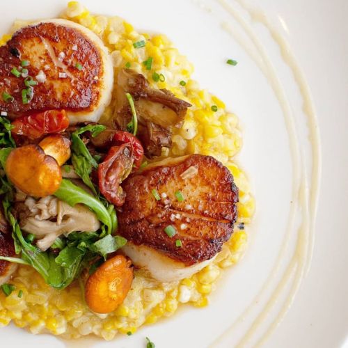 Seared scallops on a bed of creamy risotto, garnished with greens, cherry tomatoes, and mushrooms.