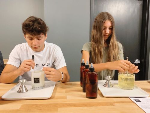 Two young people are mixing ingredients in containers on a table, with bottles and measuring tools beside them.
