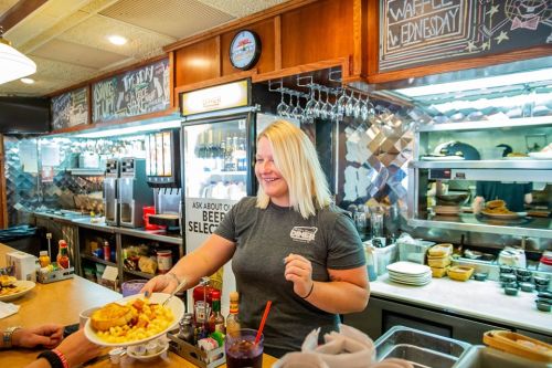 A smiling waitress serves a plate of food at a diner, with a cozy kitchen and chalkboard menu in the background.
