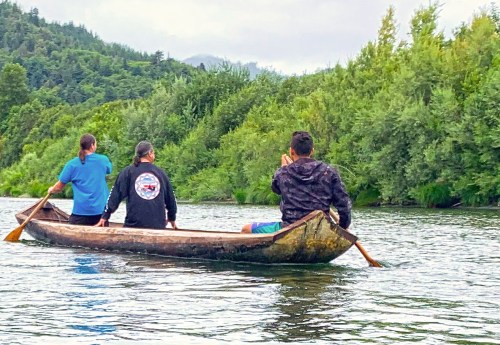 Three people paddle a wooden canoe on a calm river, surrounded by lush greenery and distant mountains.
