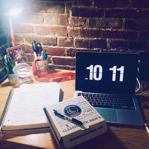 A study desk with a laptop displaying the time, notes, a cookie box, and a lamp, set against a brick wall.