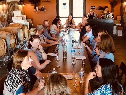 A group of people seated at a long table in a rustic venue, engaged in conversation with wine glasses in front of them.