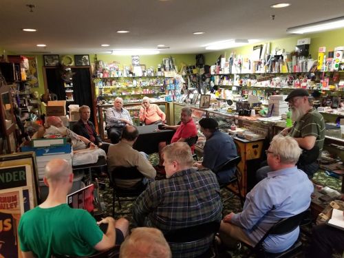 A group of people seated in a store filled with various collectibles and items, engaged in conversation.