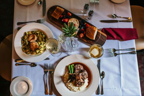 A table set with two gourmet dishes: pasta with scallops and a steak with asparagus, alongside bread and a drink.