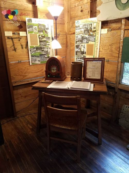 A vintage wooden desk with a chair, an old radio, and a framed document in a rustic room with wooden walls.