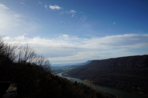 A scenic view of a river winding through valleys, framed by trees and mountains under a blue sky with scattered clouds.