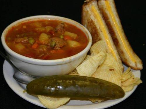 A bowl of hearty soup with vegetables and meat, served with a grilled cheese sandwich, potato chips, and a pickle.