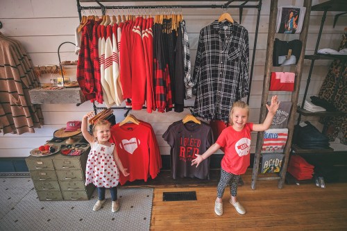 Two young girls pose in a clothing store, smiling and waving, with red and black apparel displayed on racks behind them.