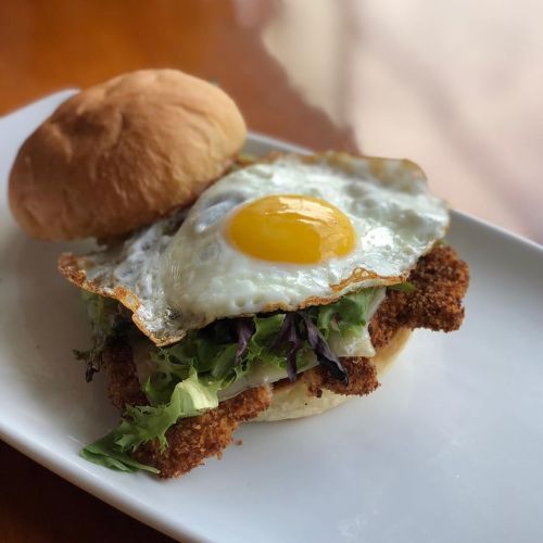 A crispy chicken sandwich topped with a fried egg and fresh greens on a soft bun, served on a white plate.
