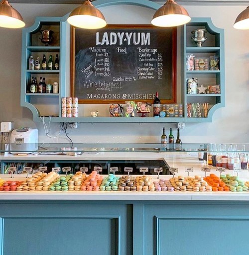 A colorful display of macarons on a counter at Lady Yum, with a chalkboard menu and shelves of treats in the background.