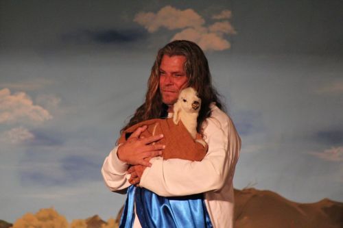 A man with long hair holds a small dog wrapped in a blanket against a scenic backdrop.