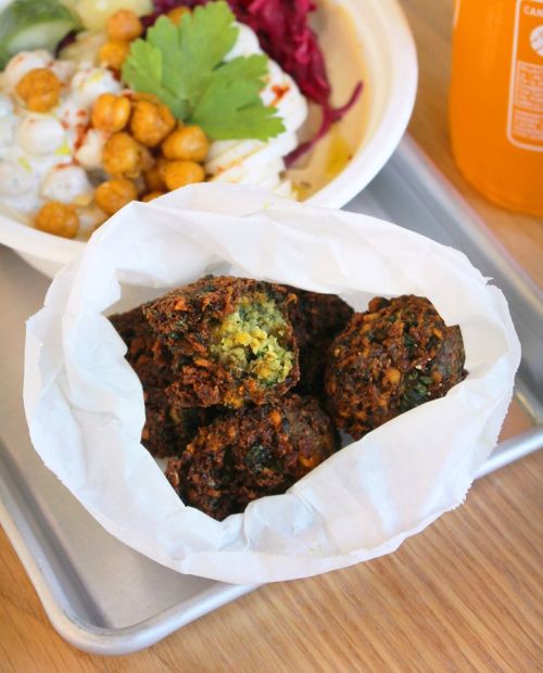 A paper container holds several golden-brown falafel balls, with a colorful salad in the background.