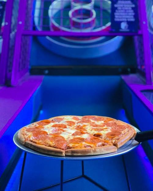A pepperoni pizza on a serving tray in a colorful arcade setting with game machines in the background.