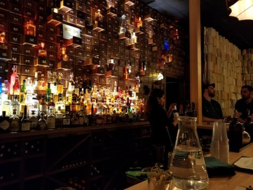 A bar scene with a wall of illuminated liquor bottles and patrons enjoying drinks at the counter.