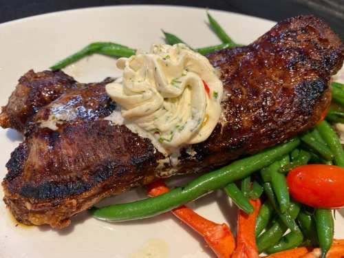 A grilled steak topped with herb butter, served on a bed of green beans and cherry tomatoes.