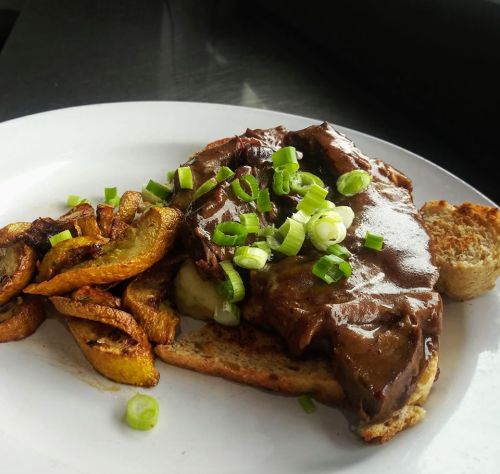 A plate featuring a hearty sandwich with beef, gravy, and green onions, accompanied by crispy fried plantains.