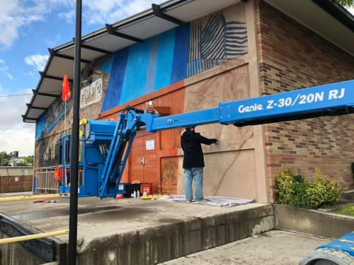 A worker on a blue lift painting a mural on a brick building with a colorful design.