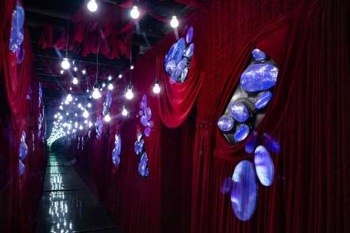 A dimly lit corridor with red drapes and glowing blue shapes on the walls, illuminated by hanging light bulbs.
