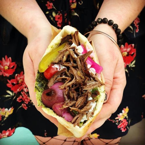A person holds a delicious wrap filled with shredded meat, pickles, onions, and colorful toppings, surrounded by a floral background.