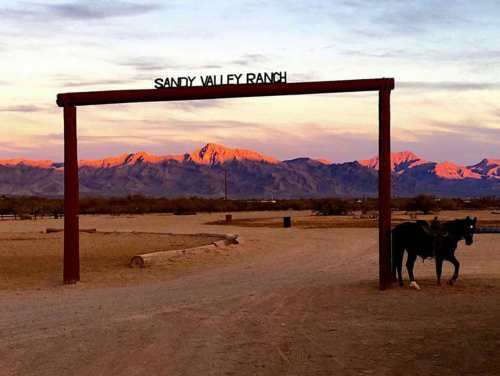 A horse stands near the entrance of Sandy Valley Ranch, with mountains bathed in sunset light in the background.