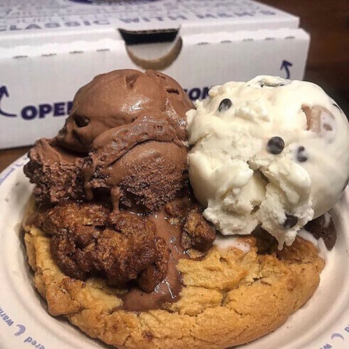 A warm cookie topped with chocolate and vanilla ice cream, garnished with chocolate chips and cookie pieces.