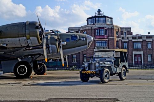 A vintage military jeep parked near a historic building and a large aircraft on a sunny day.