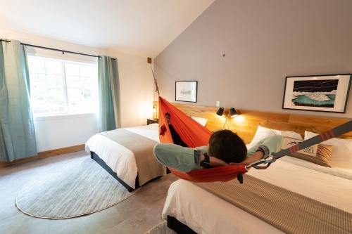 A cozy hotel room with two beds and a person relaxing in a hammock between them. Natural light from a window.