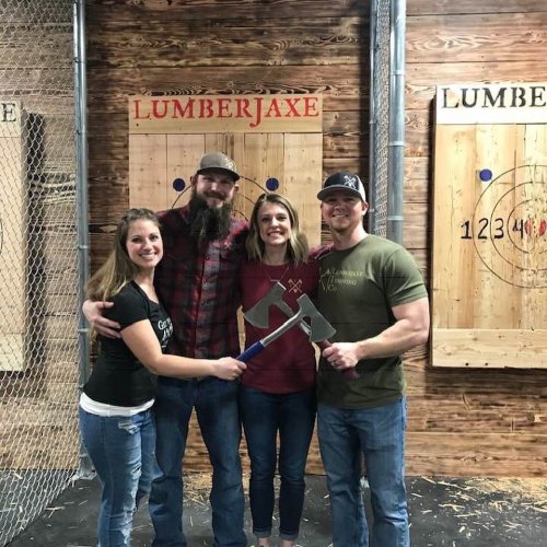 Four friends pose together at an axe-throwing venue, each holding an axe, with targets in the background.