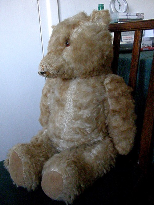 A large, plush teddy bear with a soft, light brown fur, sitting on a chair beside a wooden table.