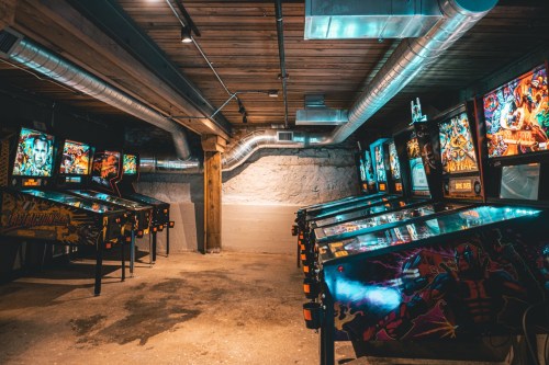 A dimly lit basement arcade featuring multiple pinball machines with colorful displays and wooden beams overhead.