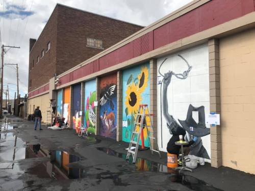 A colorful mural on a building wall, featuring various designs, with artists working and ladders nearby.