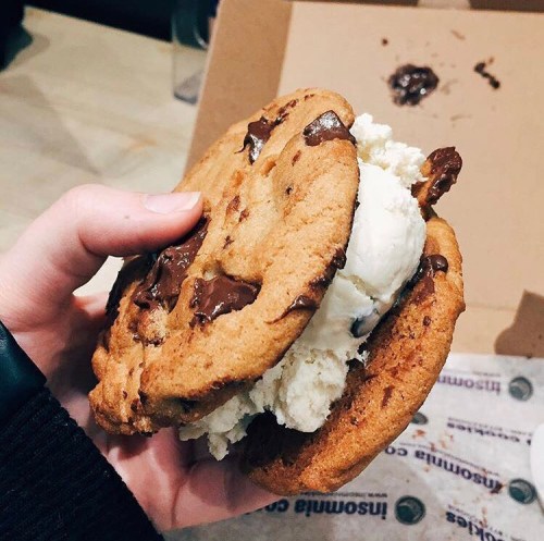 A hand holding a large ice cream sandwich made with two chocolate chip cookies and vanilla ice cream.