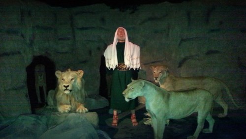 A figure in a white shawl stands among lifelike lion sculptures in a dimly lit cave setting.