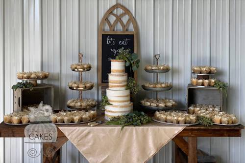 A dessert table featuring a tiered cake, cupcakes, and treats, decorated with greenery and a chalkboard sign.