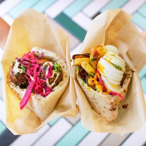 Two pita wraps filled with colorful ingredients, including falafel, pickled cabbage, and a boiled egg, served in paper.